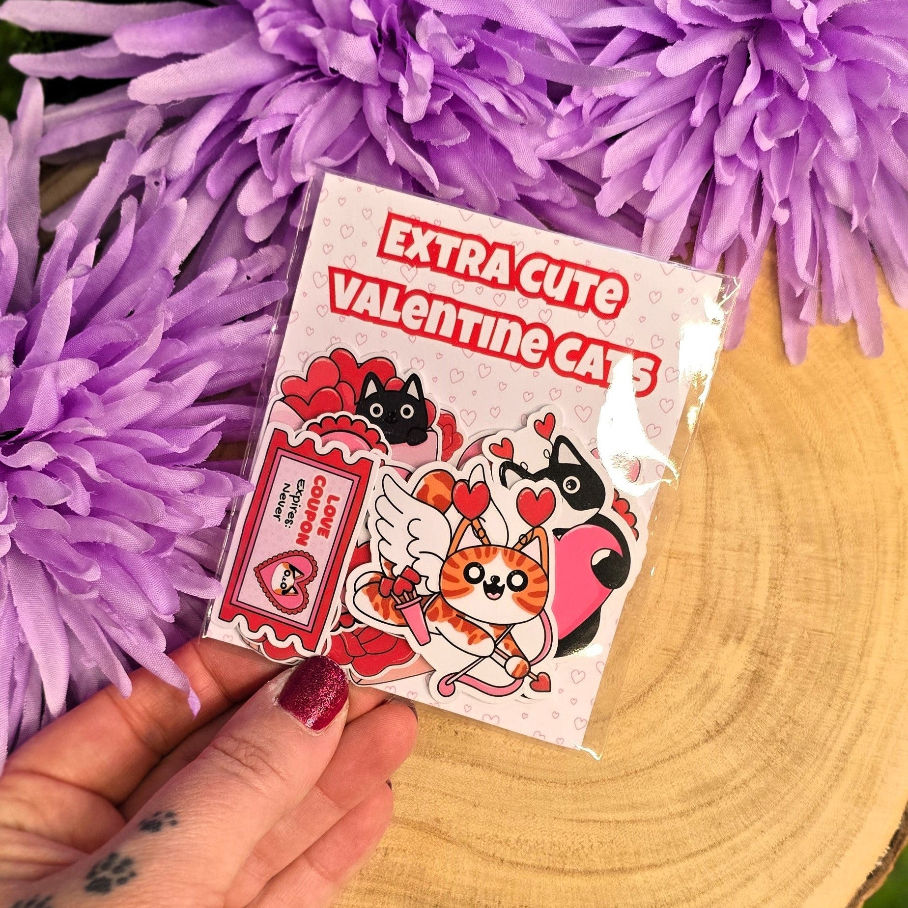Hand holding a cats sticker pack and text 'Extra Cute Valentine Cats' against a purple flower background.