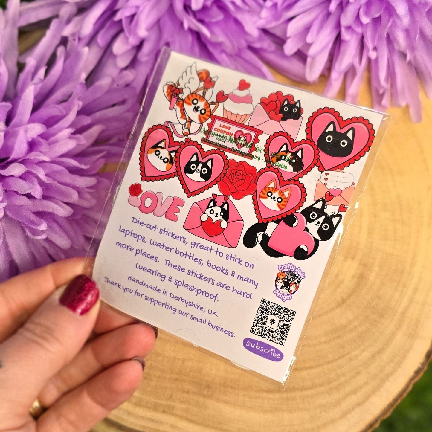 Sticker pack with cat characters and text held over a wooden surface with purple flowers in the background