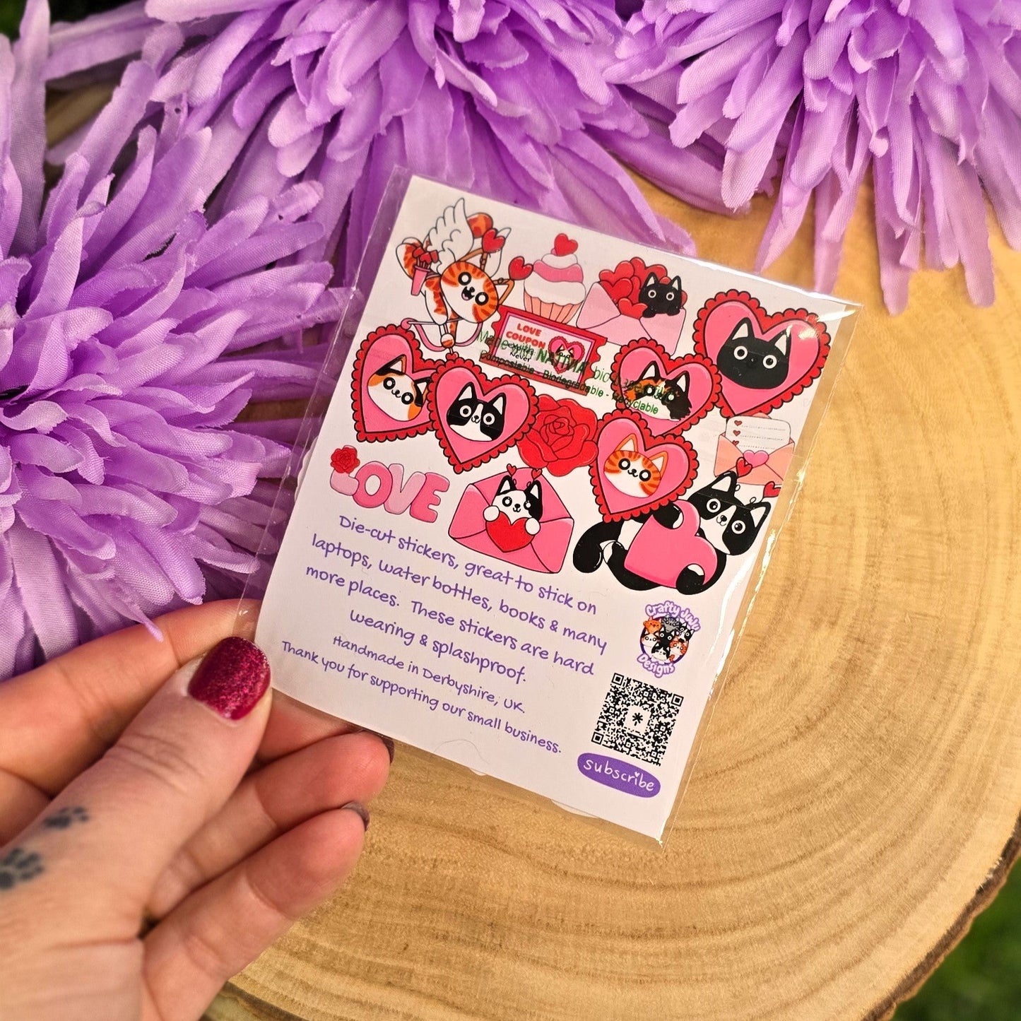 Hand holding a cat sticker pack 
 in front of purple flowers on a wooden surface