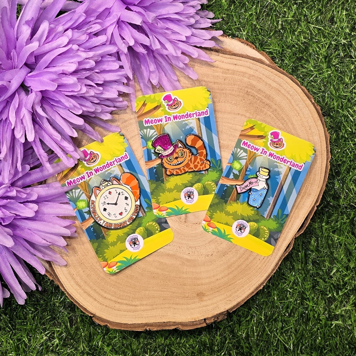 Three 'Meow in Wonderland' enamel pins on cards on a wooden surface with purple flowers and grass in the background.