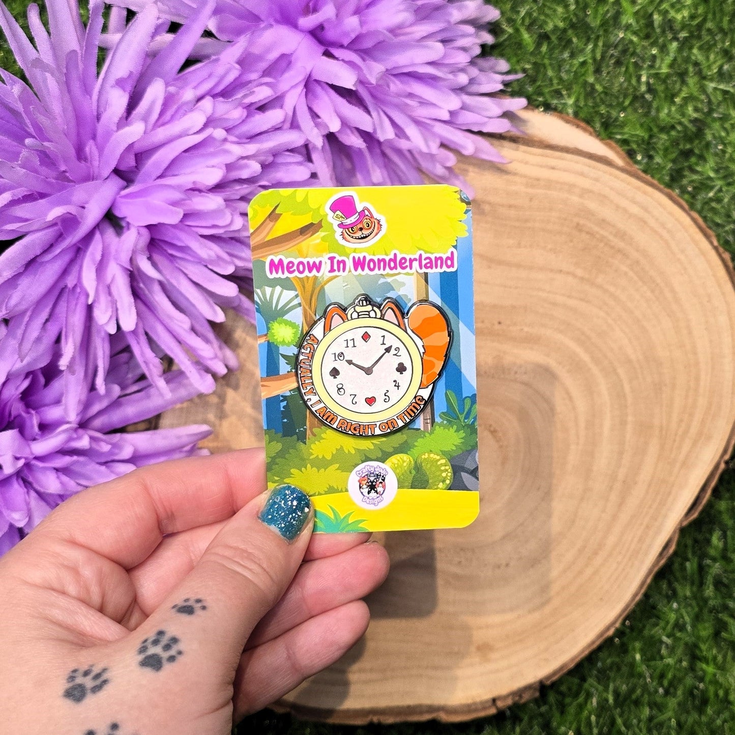 Colorful 'Meow in Wonderland' enamel pin on a card held by a hand with purple flowers and a wooden surface in the background.
