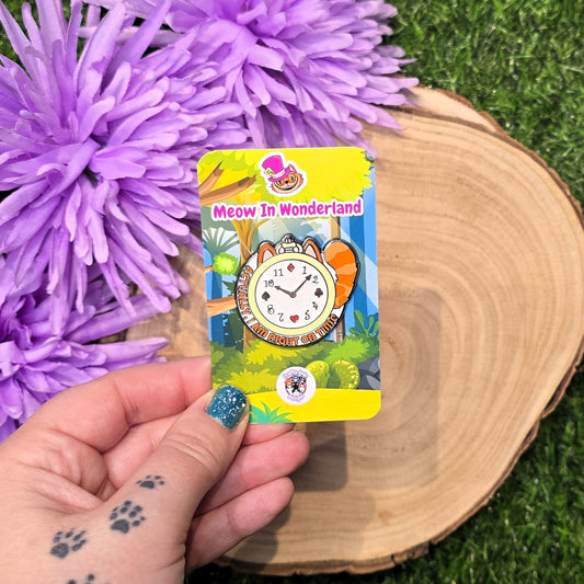 Colorful 'Meow in Wonderland' enamel pin on a card held by a hand with purple flowers and a wooden surface in the background.