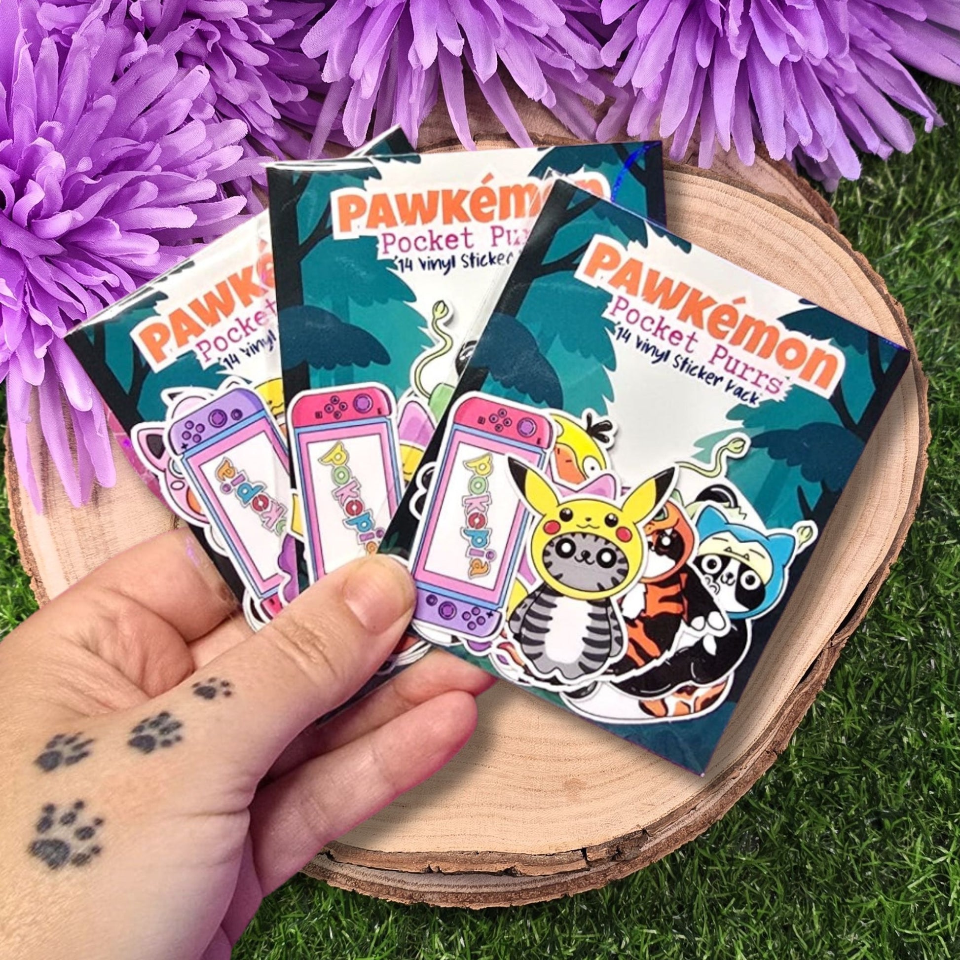 Pawkémon Pocket Purrz sticker packs held by a hand with purple flowers and grass in the background