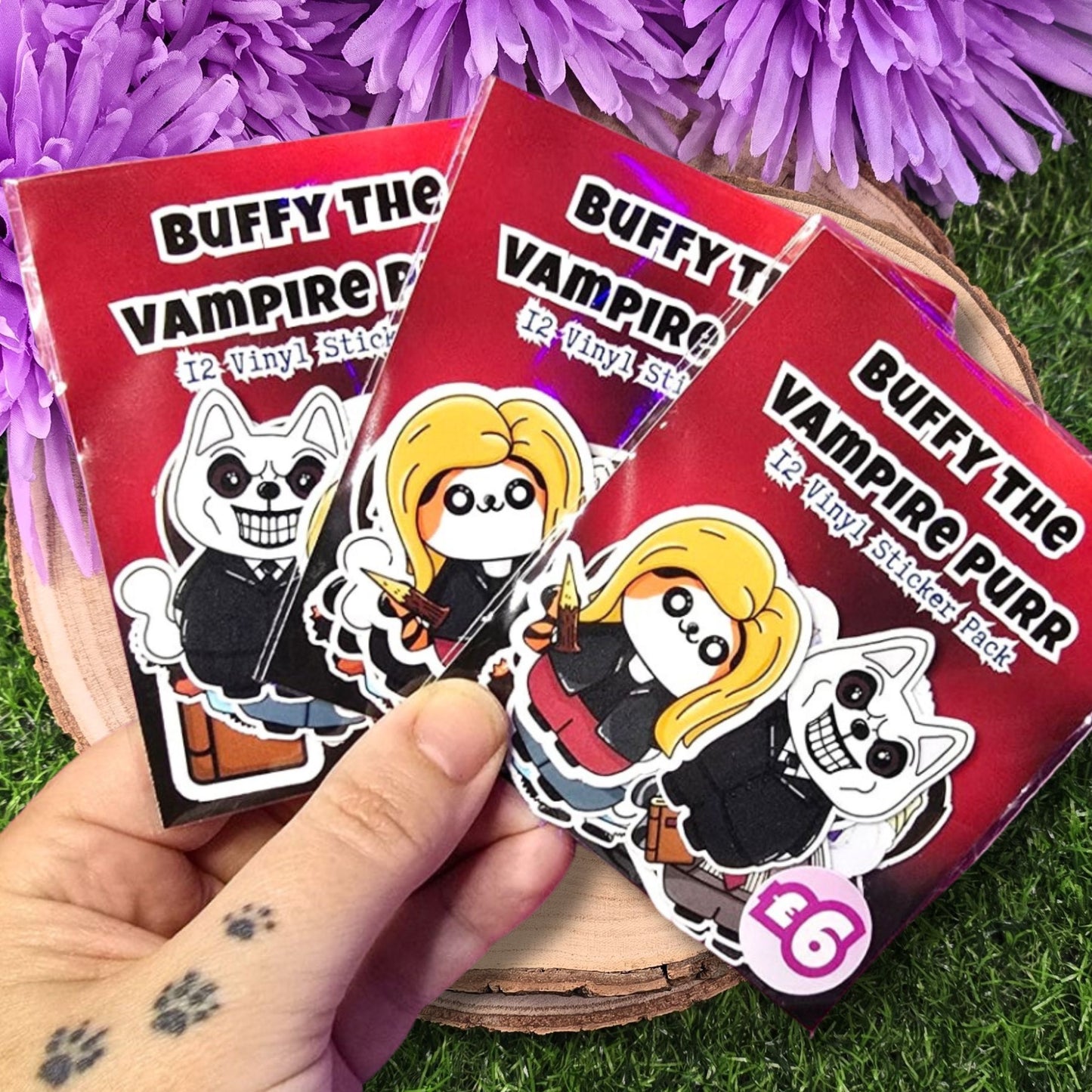 Three packs of 'Buffy the Vampire Purr' stickers held by a hand on grass with purple flowers.