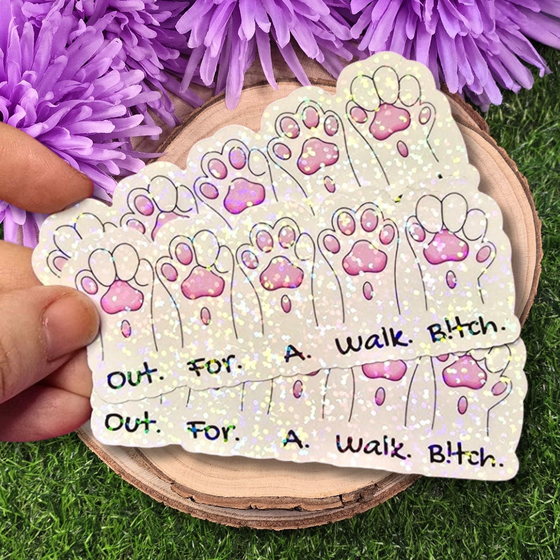 Holographic sticker with paw prints and text held over purple flowers and grass
