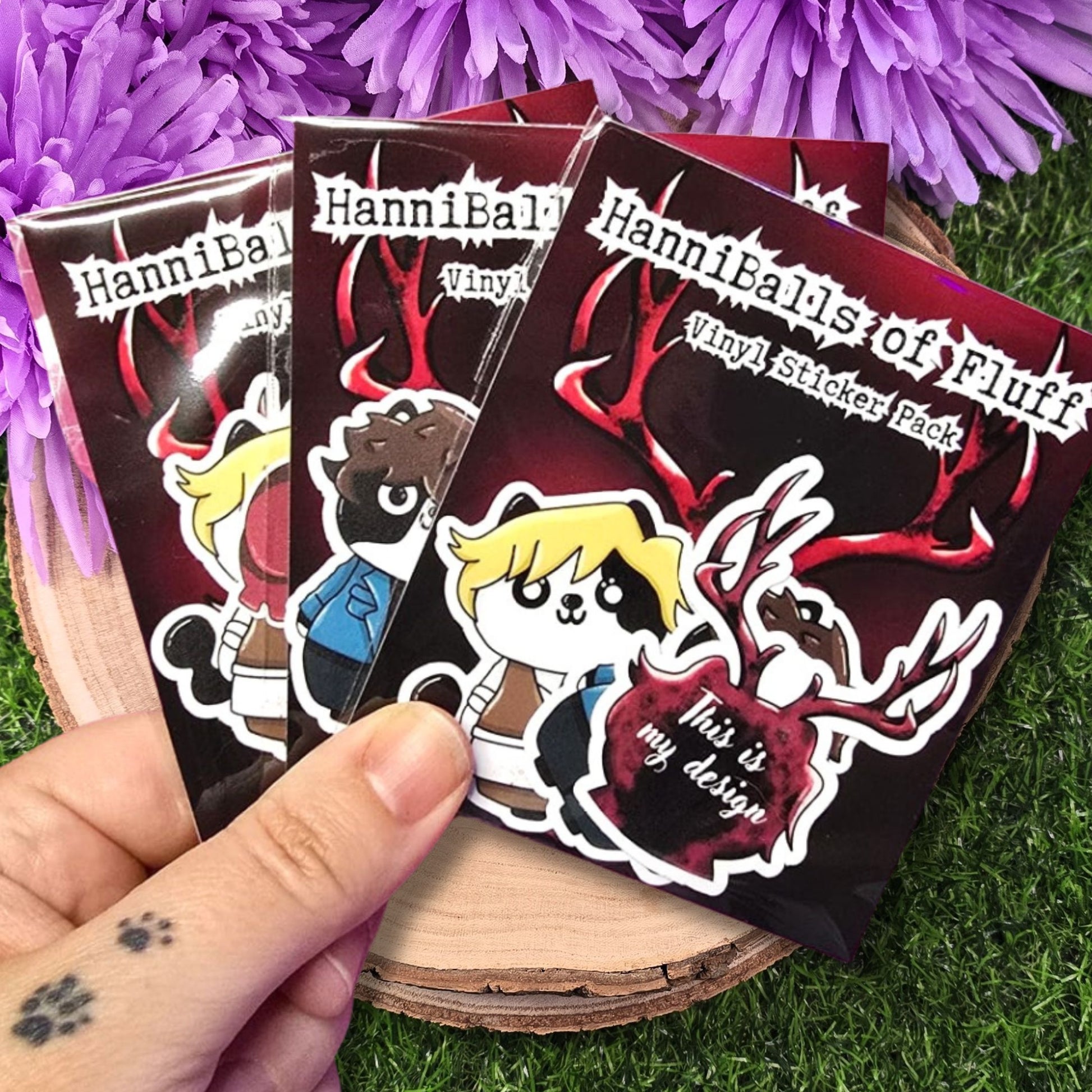 Hannibal Balls of Fluff vinyl sticker pack held by a hand with purple flowers and grass in the background