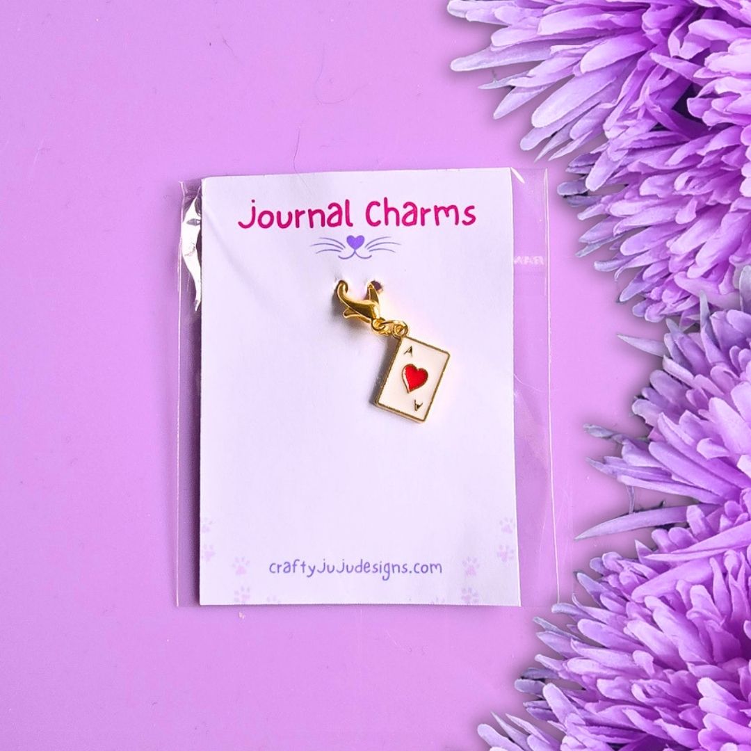 A gold playing card charm with a red heart, attached to a white display card labeled 'Journal Charms' and 'craftyJuJuddesigns.com', photographed on a purple background with purple flowers along the right edge.