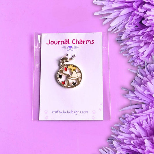 A round clock-shaped journal charm with a silver-toned metal frame and colorful details, displayed on a white card labeled 'Journal Charms' by Crafty Ju Ju Designs, set against a purple background with purple flowers on the side.