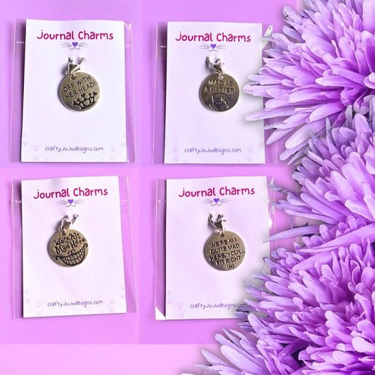 Four round metal journal charms on white display cards, arranged in a 2x2 grid on a purple background with purple flowers along the right side. The cards are branded 'Journal Charms' and show the website crafty-jujulesigns.