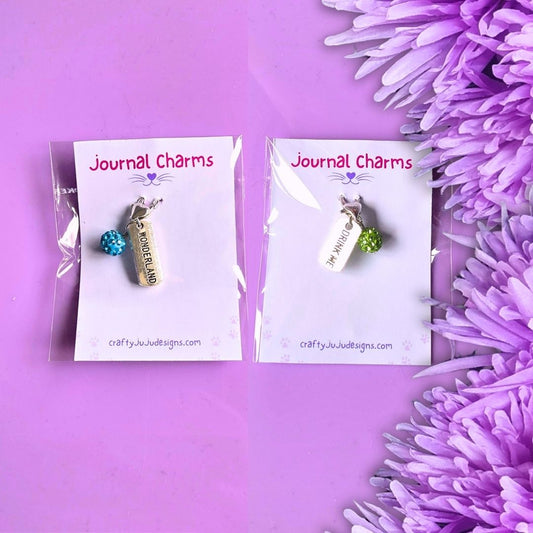 Two small journal charms on white display cards, photographed on a purple background with purple flowers along the right side. The left charm is a silver bar with a blue bead and the text 'Wonderland'; the right charm is a tag with a green bead and the text 'Drink Me'.
