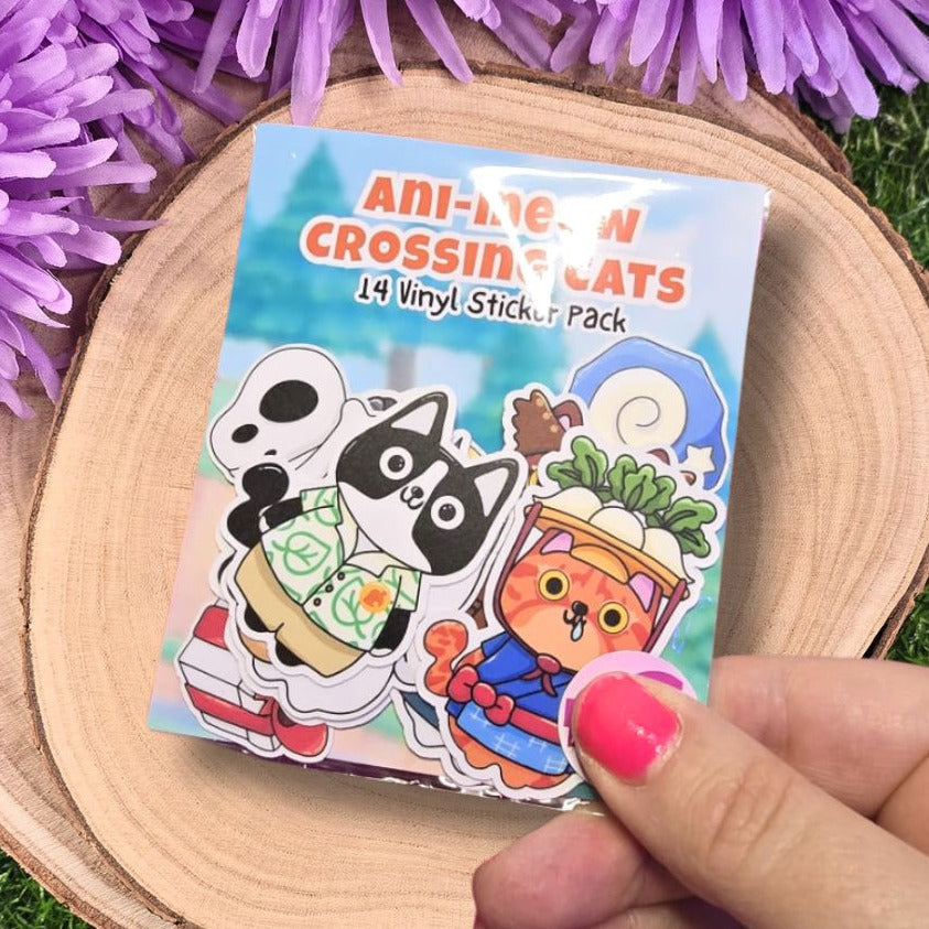 Pack of 'Anime Crossing Cats' vinyl stickers held by a hand with pink nail polish, surrounded by purple flowers on a wooden surface.  Meet all 14 island residents!