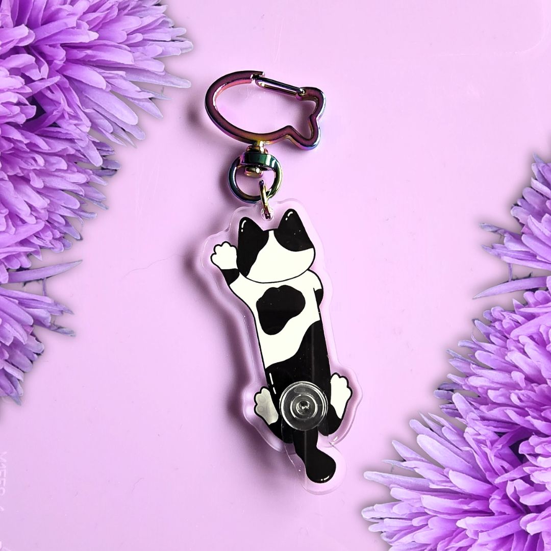 back of a black and white cow print cat keyring with moveable tail and fish shaped keyring attachment. laid on a purple background with purple flowers