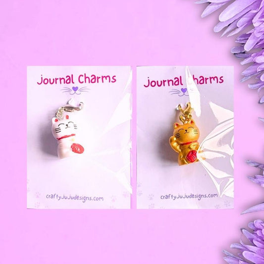 Two small cat charms on white display cards against a purple background, with purple flowers along the right side. The cards read 'Journal Charms' and show a lucky white cat charm and a gold lucky cat charm, each with a red bell.