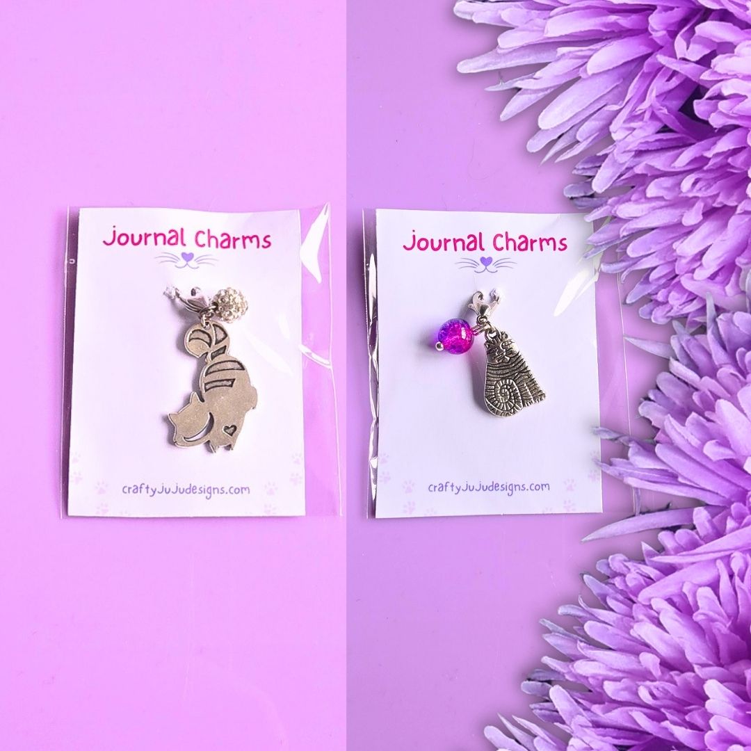 Two metal journal charms on white display cards against a lavender background, with purple flowers along the right side. The left charm is silver-toned and shaped like a cat, and the right charm is silver-toned with a pink gem accent and cat charm.