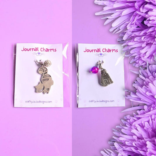 Two metal journal charms on white display cards against a lavender background, with purple flowers along the right side. The left charm is silver-toned and shaped like a cat, and the right charm is silver-toned with a pink gem accent and cat charm.