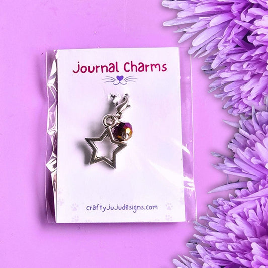 A silver metal journal charm featuring an open star and a small bead, presented on a white card labelled 'Journal Charms' by craftyJuJudesigns.com, set against a purple background with purple flowers.