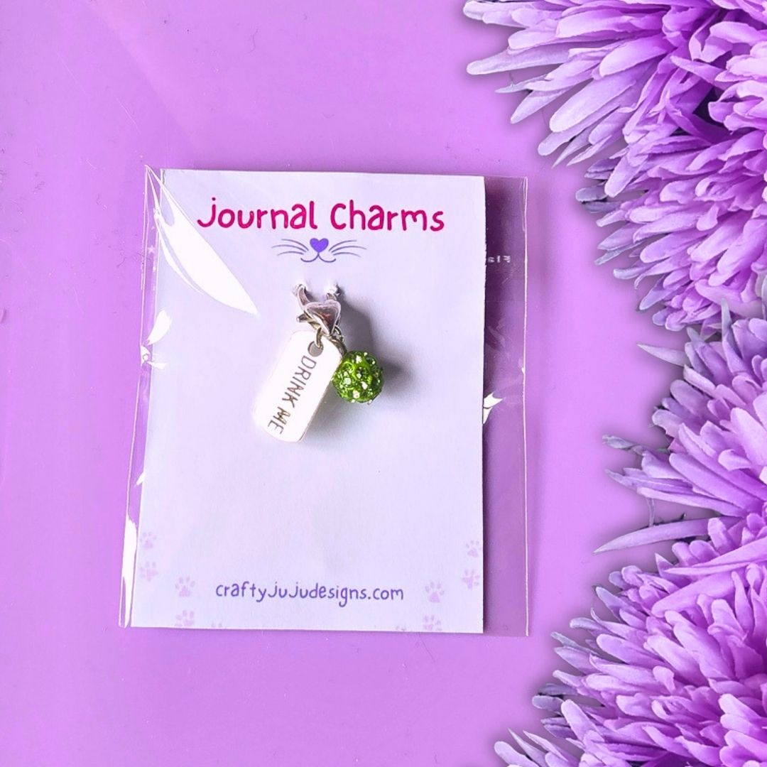 A silver journal charm with a green rhinestone apple and the word 'Drink Me' on a white tag, displayed on a branded card over a purple background with purple flowers.