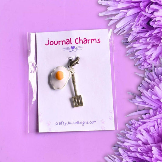 A charm shaped like a fried egg with a small spatula hanging from it, displayed on a white card labeled 'Journal Charms' against a lavender background with purple flowers.