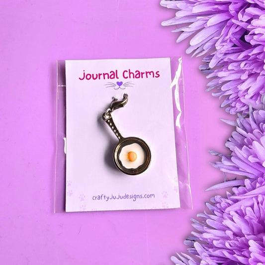 A small frying pan charm with a yellow-orange egg inside, attached to a white display card labeled 'Journal Charms' and photographed on a pink-purple background with purple flowers at the side.