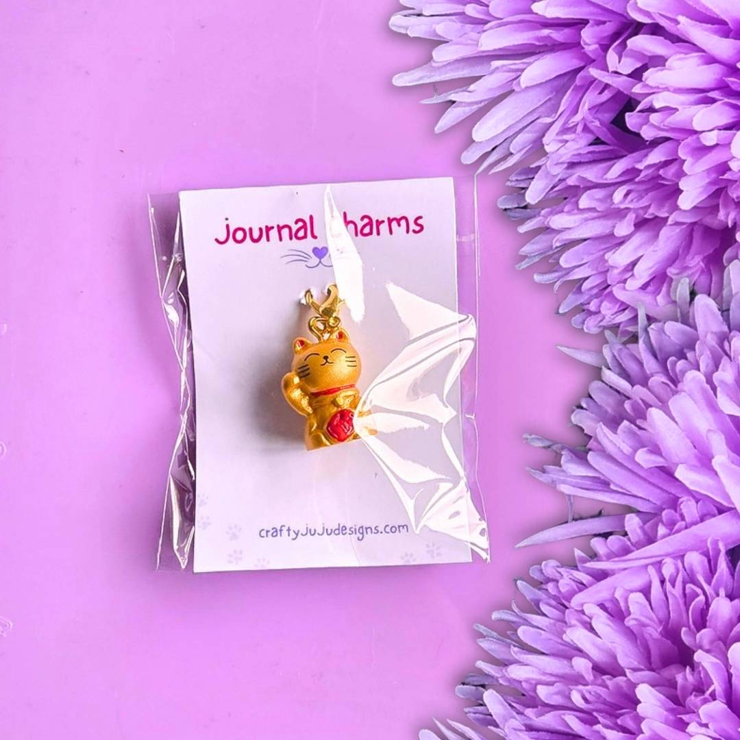 A gold-toned Maneki-neko cat charm with a red pendant, displayed on a white card labeled 'Journal charms' inside a clear plastic sleeve, set against a purple background with purple flowers.