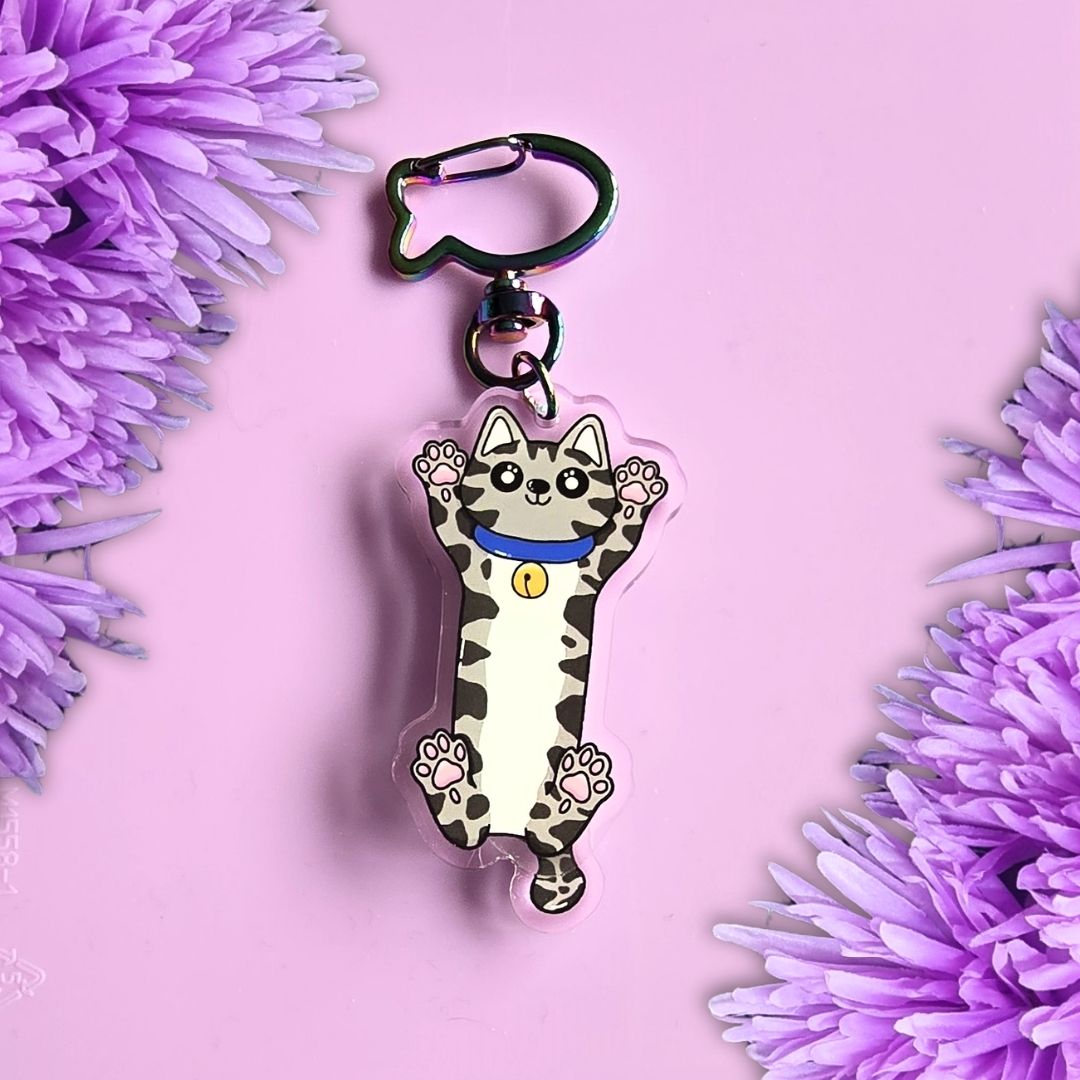 grey tabby cat stretched out acrylic keyring with moving tail and fish shaped key ring clasp. All laid on a purple background with purple flowers around the edges
