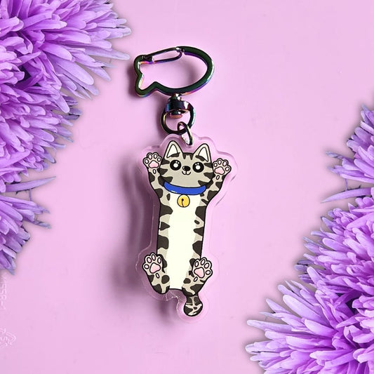 grey tabby cat stretched out acrylic keyring with moving tail and fish shaped key ring clasp. All laid on a purple background with purple flowers around the edges
