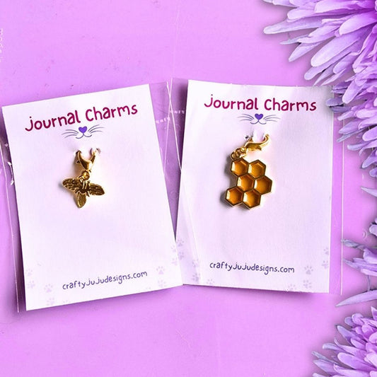 Two gold bee-themed journal charms on white display cards, styled with purple flowers on a lavender background. The left card shows a single bee charm, and the right card shows a honeycomb bee charm.