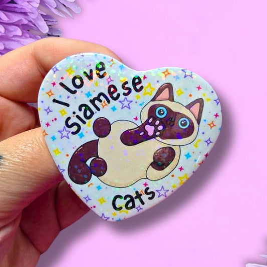 A hand holds a heart-shaped pin badge featuring a cartoon Siamese cat and the text 'Love Siamese Cats' against a light background with colorful stars. The hand is positioned on the left, with purple flowers framing the top-left and bottom-right corners, set against a solid light pink background.
