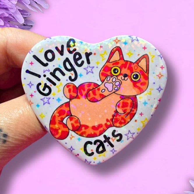 A hand holds a heart-shaped pin featuring a colorful illustration of an orange ginger cat with the text 'I love ginger cats' on a light blue starry background. The pin is set against a pink backdrop decorated with purple flowers.
