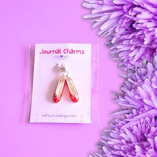 A pair of red ballet flat charms with silver trim on a clear display card labeled 'Journal Charms', photographed on a pink background with purple flowers along the right side.
