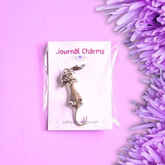 A silver-toned metal cat charm displayed on a white backing card labeled 'Journal Charms' and 'craftycharms.com', photographed on a pink background with purple flowers along the right side.