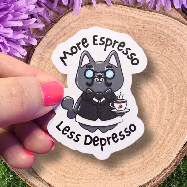 Sticker with a cat design and text 'More Espresso Less Depresso' held over a wooden surface with purple flowers.