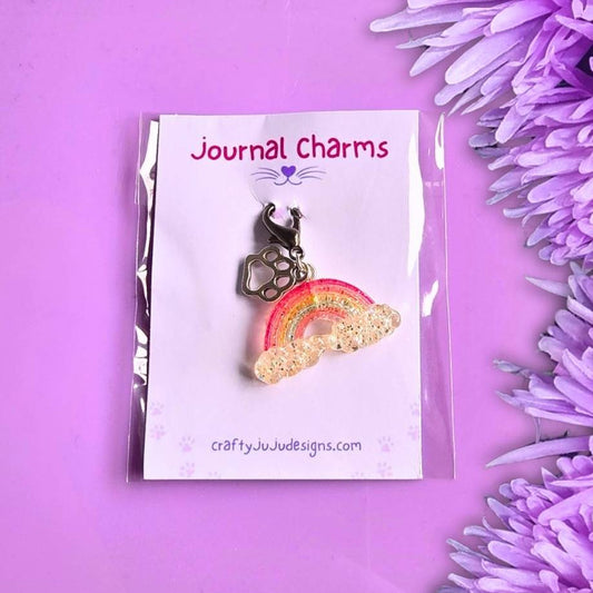 Over The Rainbow Bridge clip on charm is a rainbow with a paw print charm on the clip. That is inside its packaging on white card. That is sitting on a purple background with purple flowers to the right