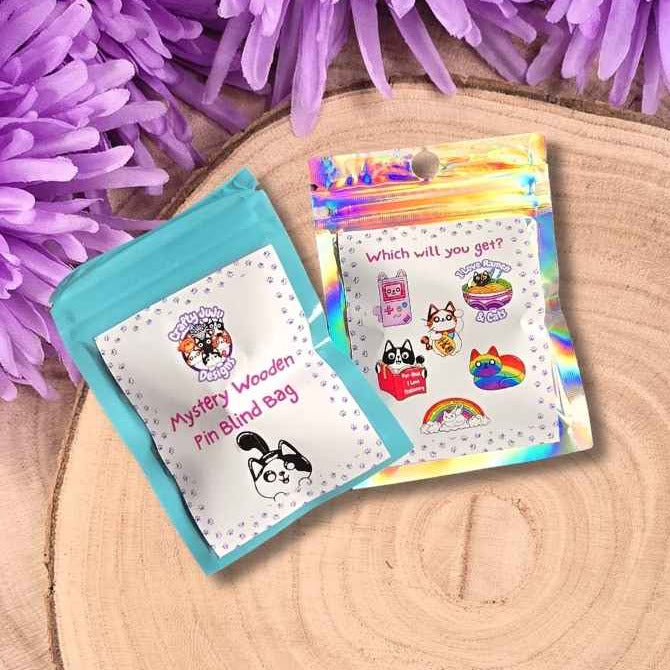 Two small pin mystery bags placed on a round wood slice, surrounded by purple flowers and green grass. One pouch is light aqua with colourful cartoon illustrations and the text 'what will you get?'.