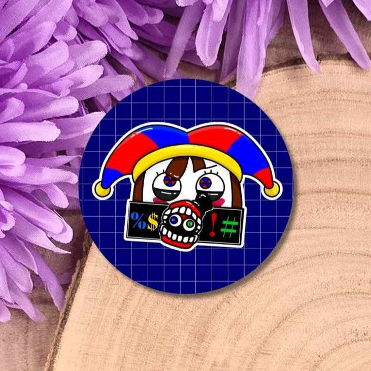A round pin badge featuring a cartoon jester character with a red, yellow, and blue hat, set against a dark blue grid background. The badge is styled on a wooden surface surrounded by purple flowers.