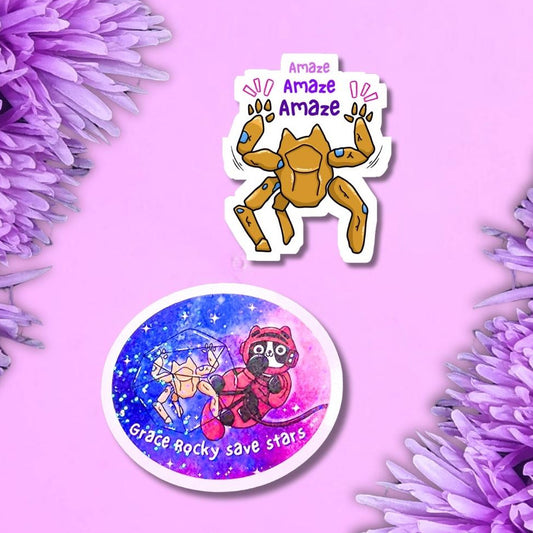 Two die-cut stickers displayed on a purple background decorated with purple flowers. The top sticker features rocky with the text 'Amaze Amaze Amaze' and the other ticker is of Rocky and Grace floating in space and the words 'Grace Rocky Save Stars'