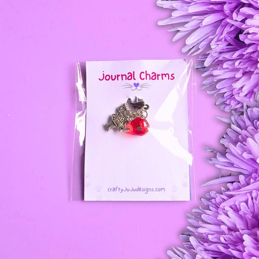 A silver-toned cat charm with a red bead accent, displayed on a white backing card labeled 'Journal Charms' by craftyJuJudedesigns.com, set against a purple background with purple flowers on the right.