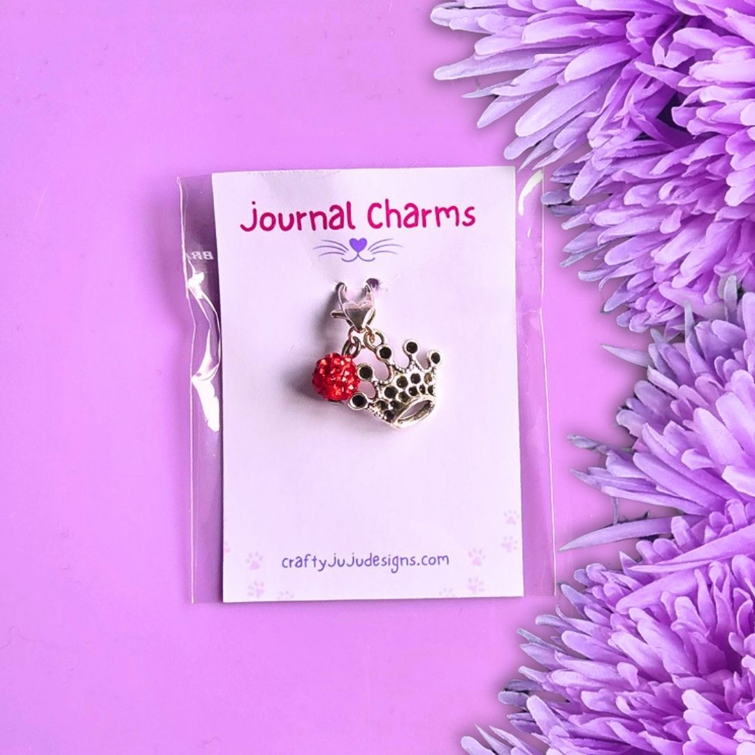 A crown charm with a red bead, displayed on a white card labeled 'Journal Charms' against a purple background with purple flowers.