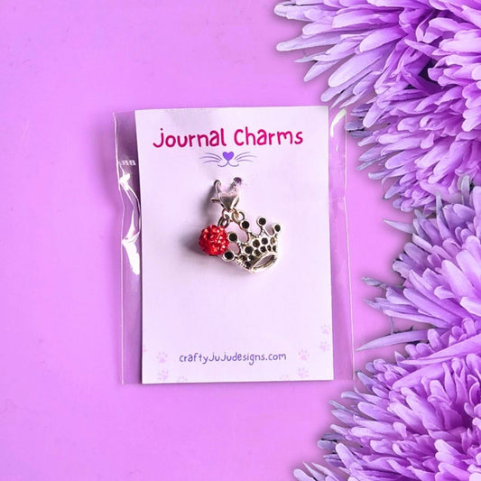 A crown charm with a red bead, displayed on a white card labeled 'Journal Charms' against a purple background with purple flowers.