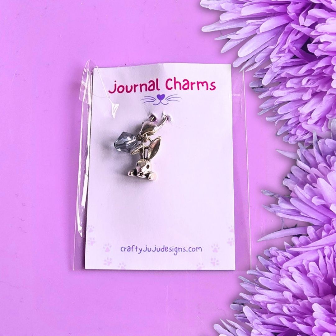A silver metal journal charm shaped like a rabbit head with a blue bead, attached to a white backing card labeled 'Journal Charms' and photographed on a purple background with purple flowers along the right side.