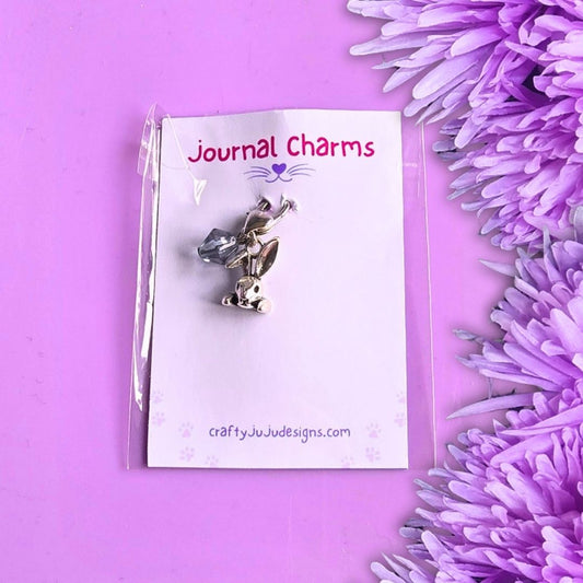 A silver metal journal charm shaped like a rabbit head with a blue bead, attached to a white backing card labeled 'Journal Charms' and photographed on a purple background with purple flowers along the right side.