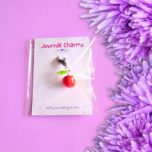 A small red apple charm with a green leaf and silver clasp, displayed on a white card labeled 'Journal Charms' against a pink background with purple flowers on the right.