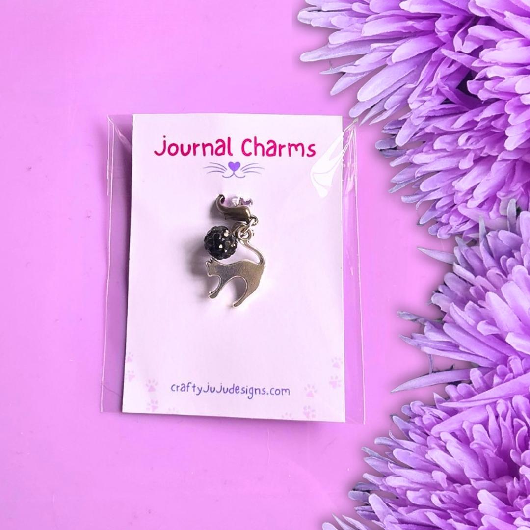 A silver Scaredy Cat charm with a black bead, on a white card labeled 'Journal Charms' by craftyjuju designs, set against a purple background with purple flowers on the right.