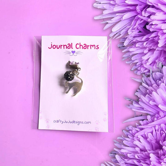 A silver Scaredy Cat charm with a black bead, on a white card labeled 'Journal Charms' by craftyjuju designs, set against a purple background with purple flowers on the right.