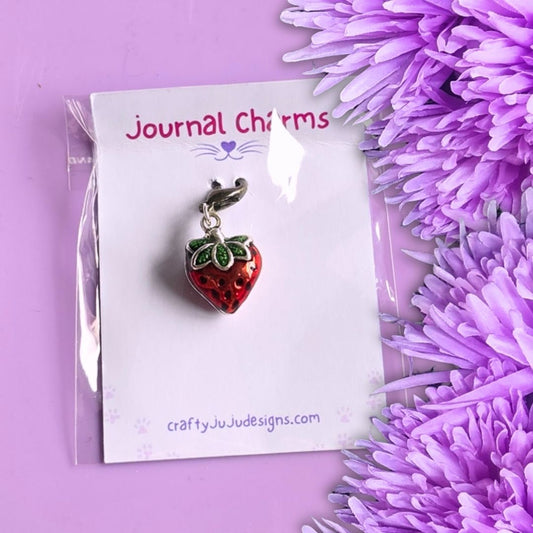 A strawberry charm with green leaves and a silver bail, displayed on a white card labeled 'Journal Charms' with purple flowers on a lavender background.
