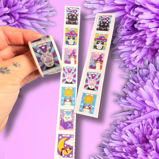 A hand holds a roll of decorative washi tape, with two unrolled strips showing colorful cat-themed illustrations on a purple floral background.