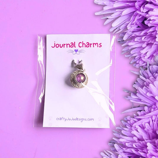A tea cup pendant charm with a purple bead inside, displayed on a white card labeled 'Journal Charms' by craftyJuJudesigns.com, set against a purple background with decorative purple flowers on the right.