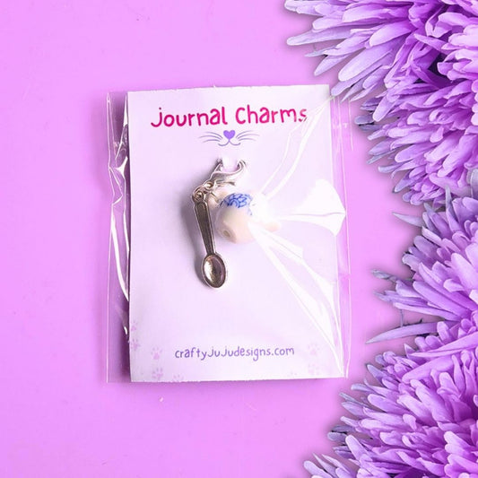 A  journal charm shaped like a teapot with blue and white floral detailing, and a spoon charm.  Attached to a white display card labeled 'Journal Charms' and photographed on a purple background with purple flowers at the side.