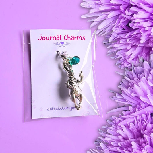 A silver-toned bunny charm with a green gemstone, displayed on a white card labeled 'Journal Charms' and photographed on a purple background with purple flowers.