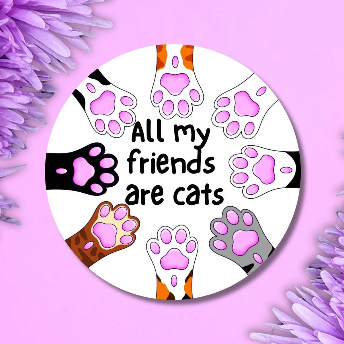 A round sticker featuring the text 'All my friends are cats' surrounded by illustrations of various cat paws in different colors on a white background, styled on a pink background with purple flowers in the corners.